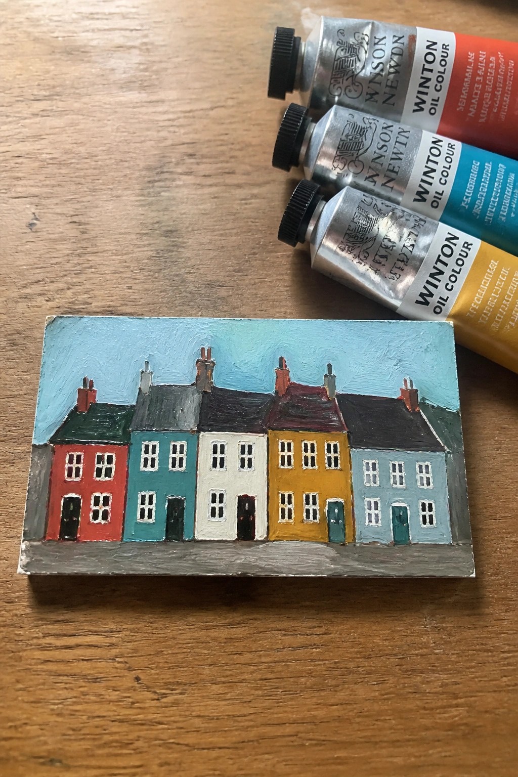 Vibrant miniature painting of colorful row houses beside oil paint tubes on wood.