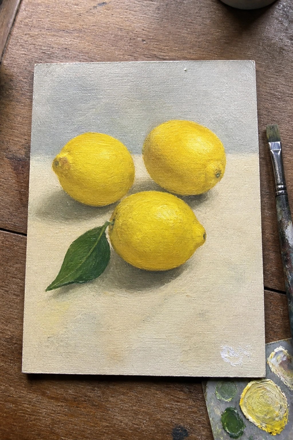 Three yellow lemons, one with green leaf, on canvas beside paintbrush and palette