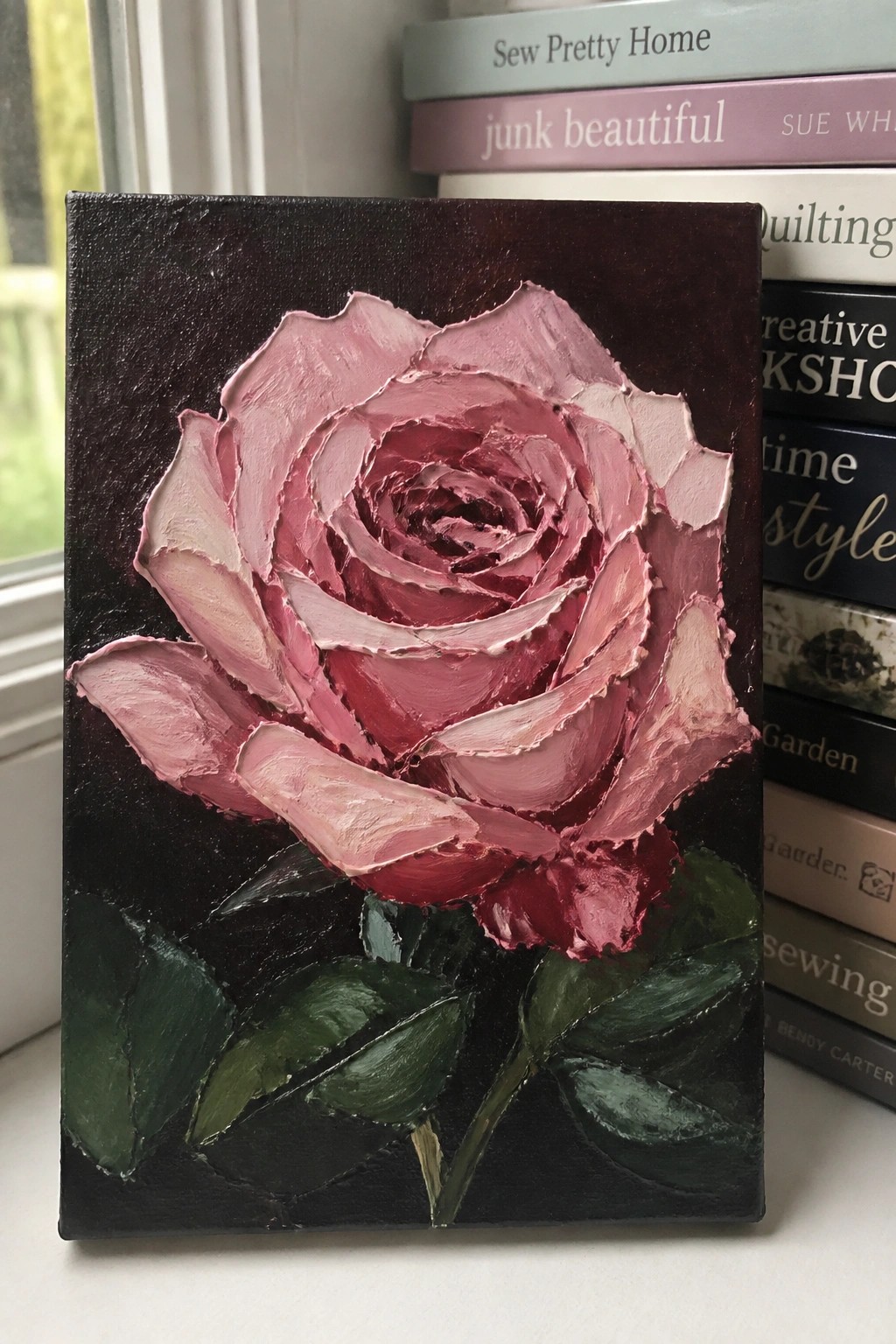 Thickly textured pink rose painting on dark canvas beside stack of books