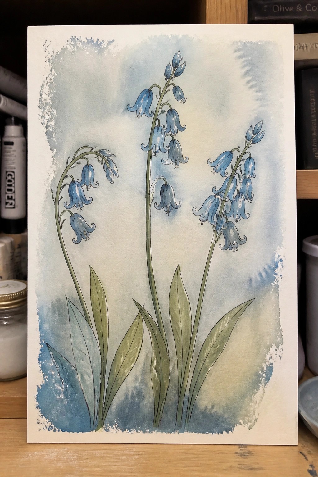 Watercolor painting of bluebell flowers with green leaves on blue-washed paper.