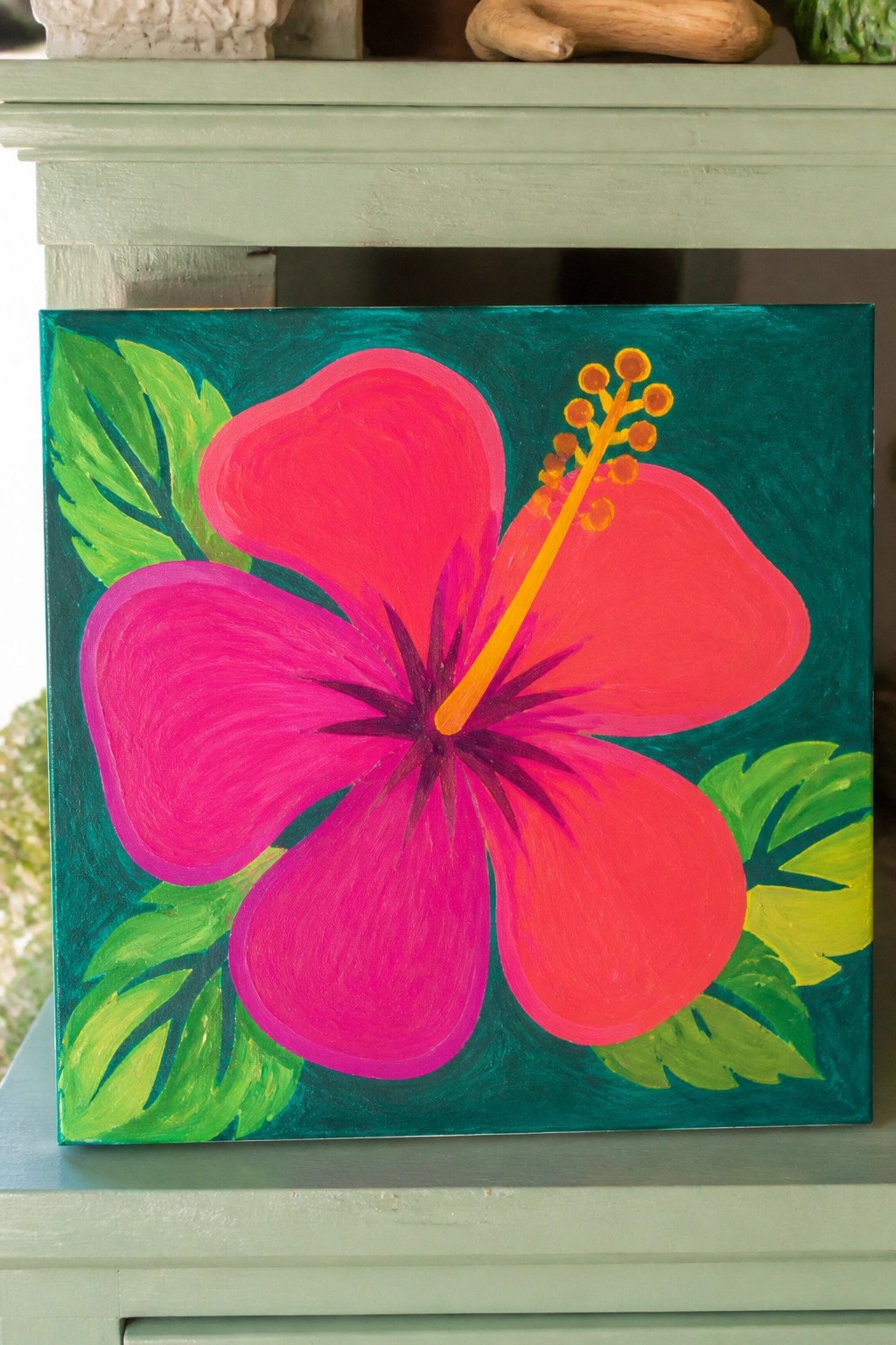 Vivid pink hibiscus flower with yellow stamens on teal canvas with green leaves