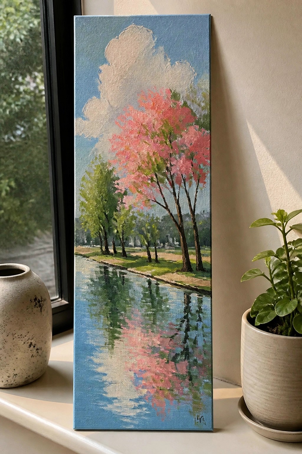 A vertical painting of pink flowering trees reflected in a blue waterway under a cloudy sky, shown on a windowsill beside a speckled vase and potted plant.
