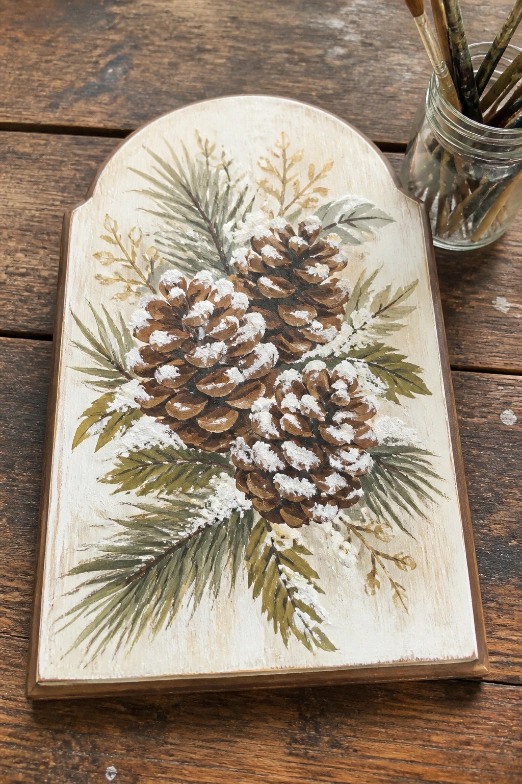 Rustic wooden plaque with snow-dusted pinecones, pine branches, and paintbrushes nearby