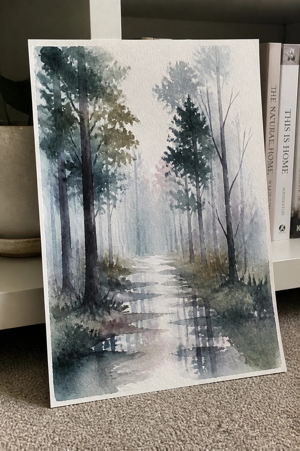 Watercolor misty forest path with tall trees and reflections beside books on shelf.