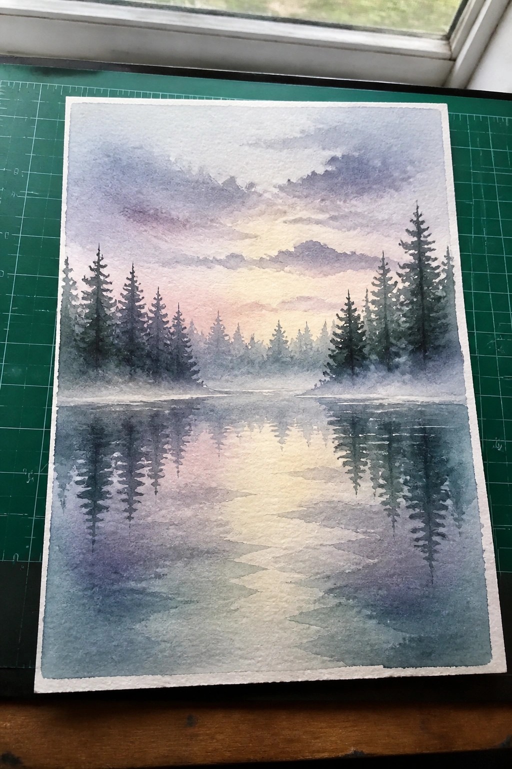 Watercolor painting of misty lake with pine trees at sunset on green cutting mat