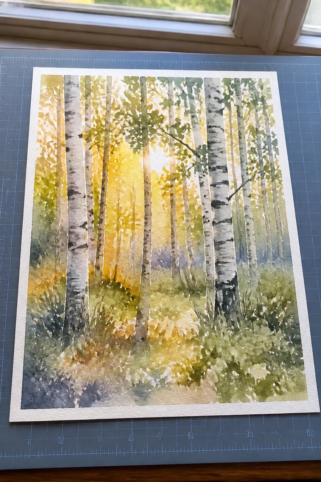Watercolor of sunlit birch forest on blue cutting mat under window