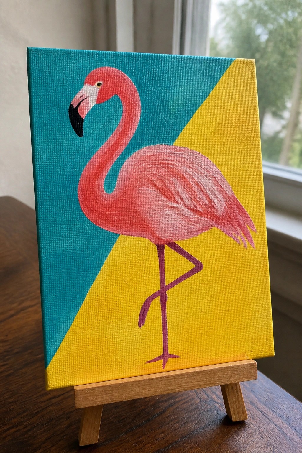 Pink flamingo painting on canvas with teal-yellow split background on wooden easel.