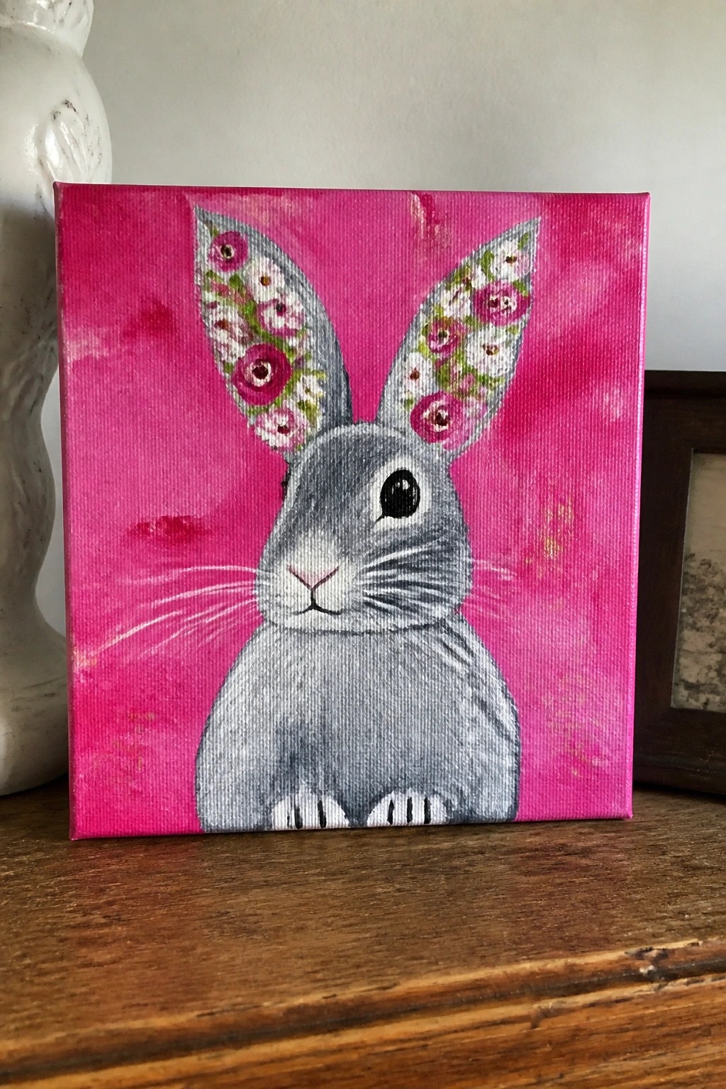 Gray rabbit with floral ears on bright pink canvas atop wooden surface