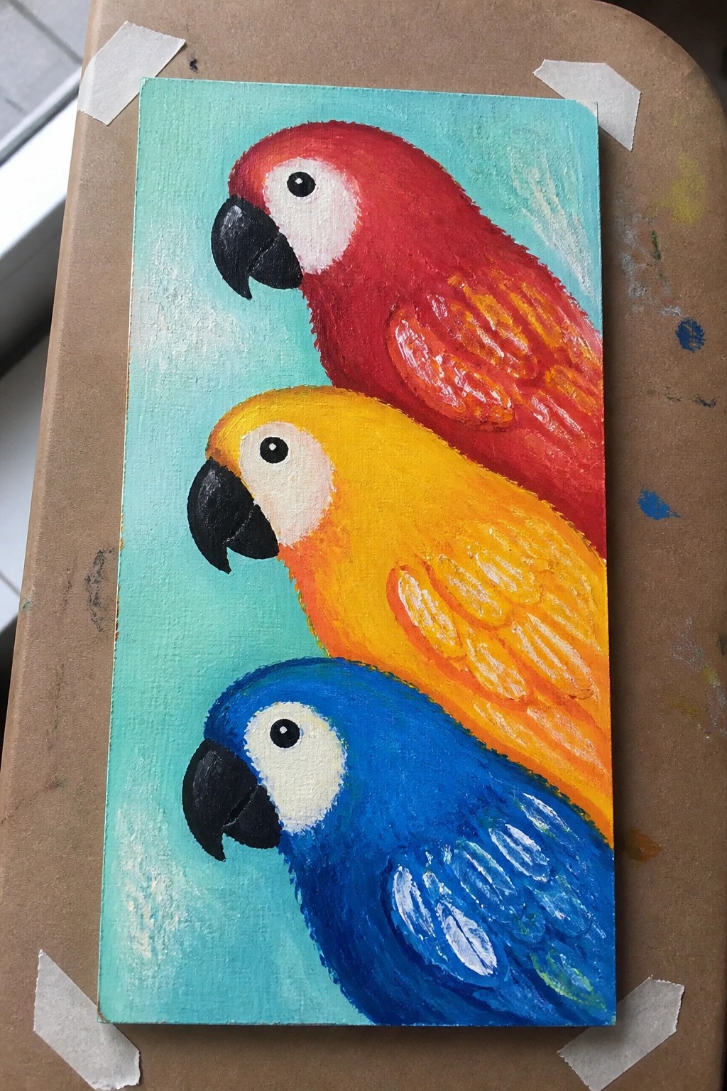 Three stacked macaws painted red, yellow, and blue on turquoise canvas