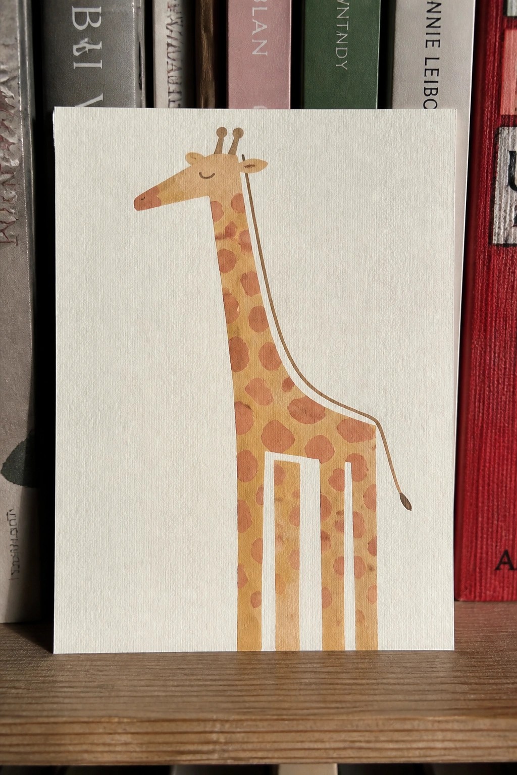 Watercolor giraffe with brown spots on white card before bookshelf of books