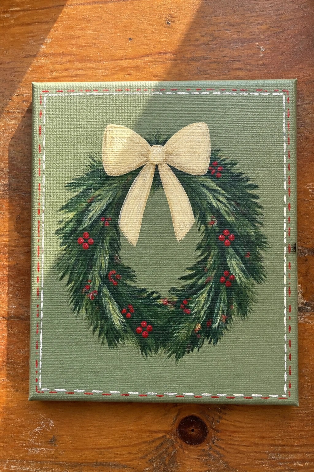 Canvas painting of Christmas wreath with green branches, red berries, and cream bow on green background.