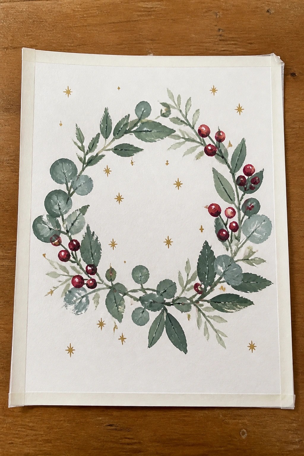 Watercolor wreath of green leaves and red berries with scattered gold stars on white paper