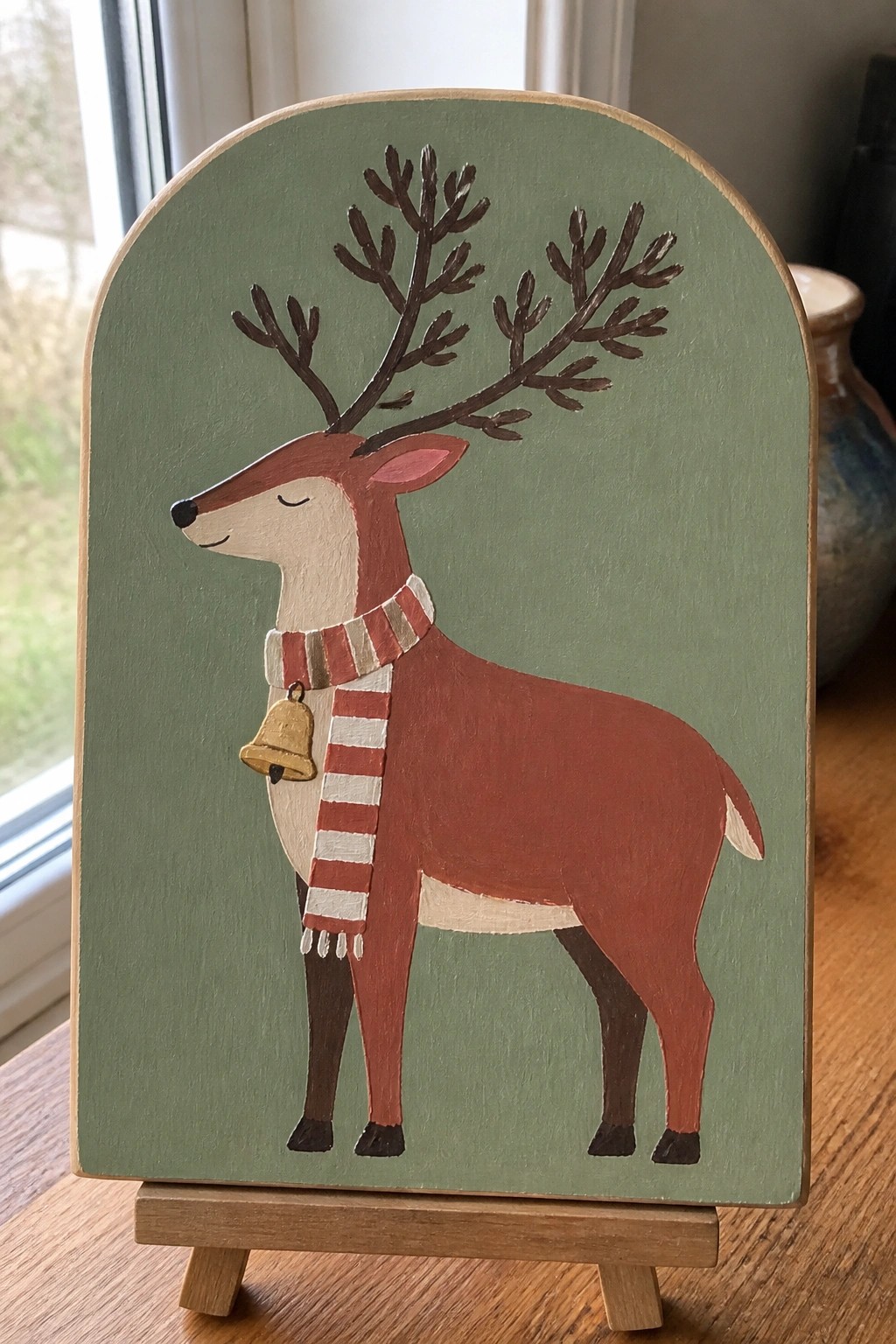 Folk art painting of reindeer with red striped scarf and bell on green arched plaque