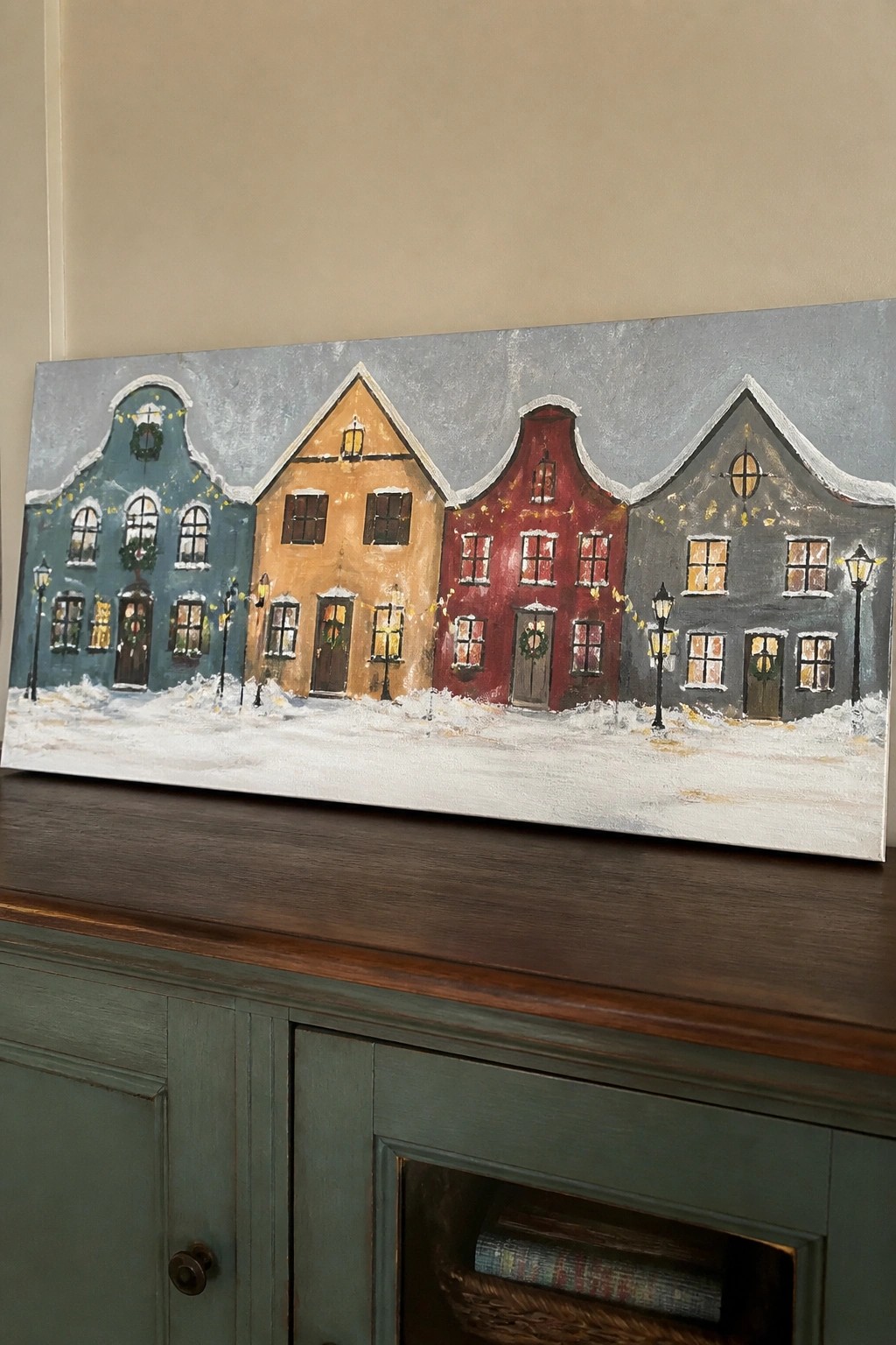 Colorful painting of four snowy houses with wreaths above green wooden cabinet.