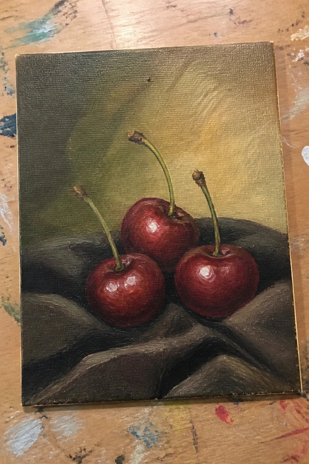 Three glossy red cherries with stems on dark wrinkled fabric, painted in oils