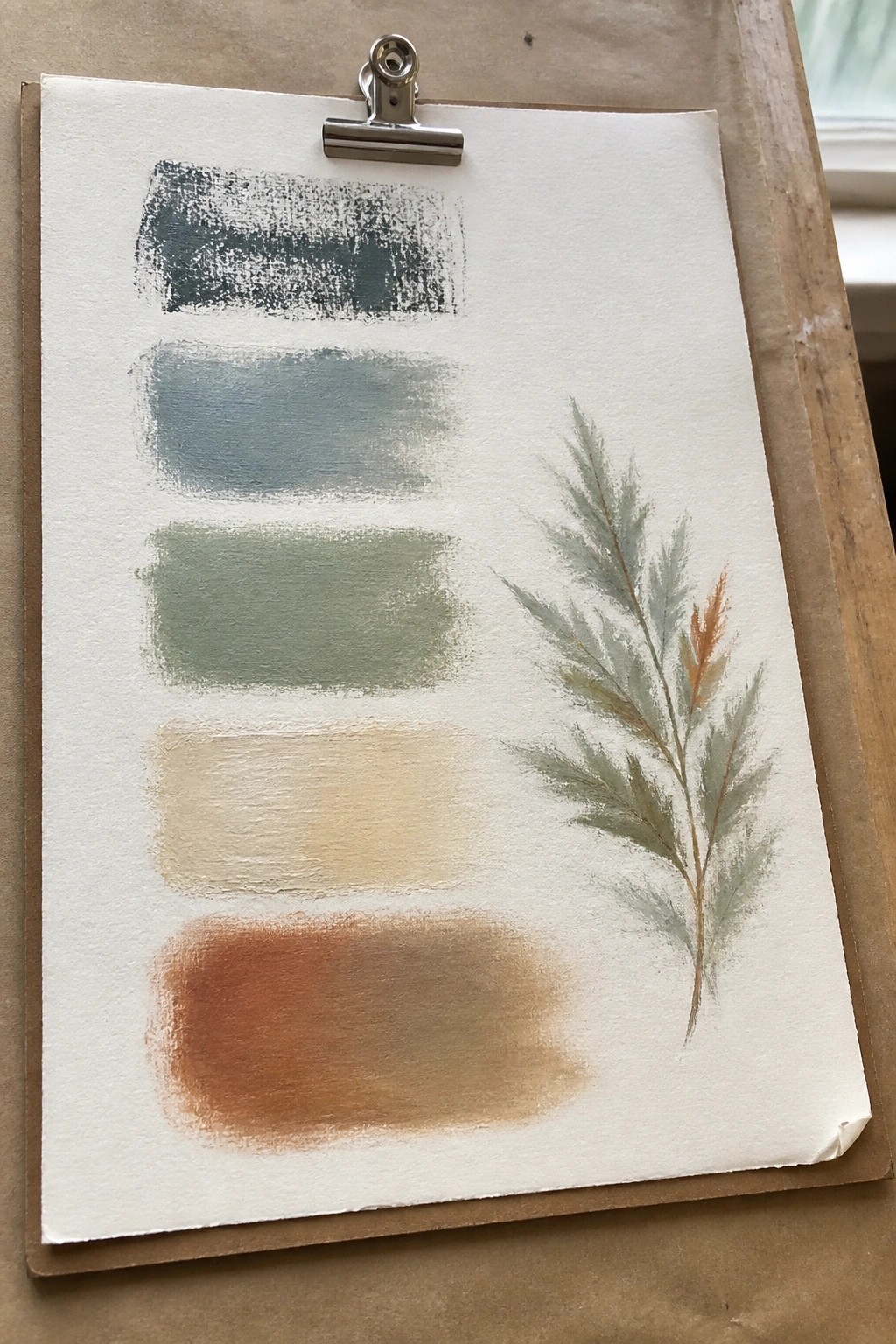 A sheet of paper on a clipboard shows five horizontal paint swatches from dark teal to rusty brown next to a painted plant sprig with muted green and brown foliage.