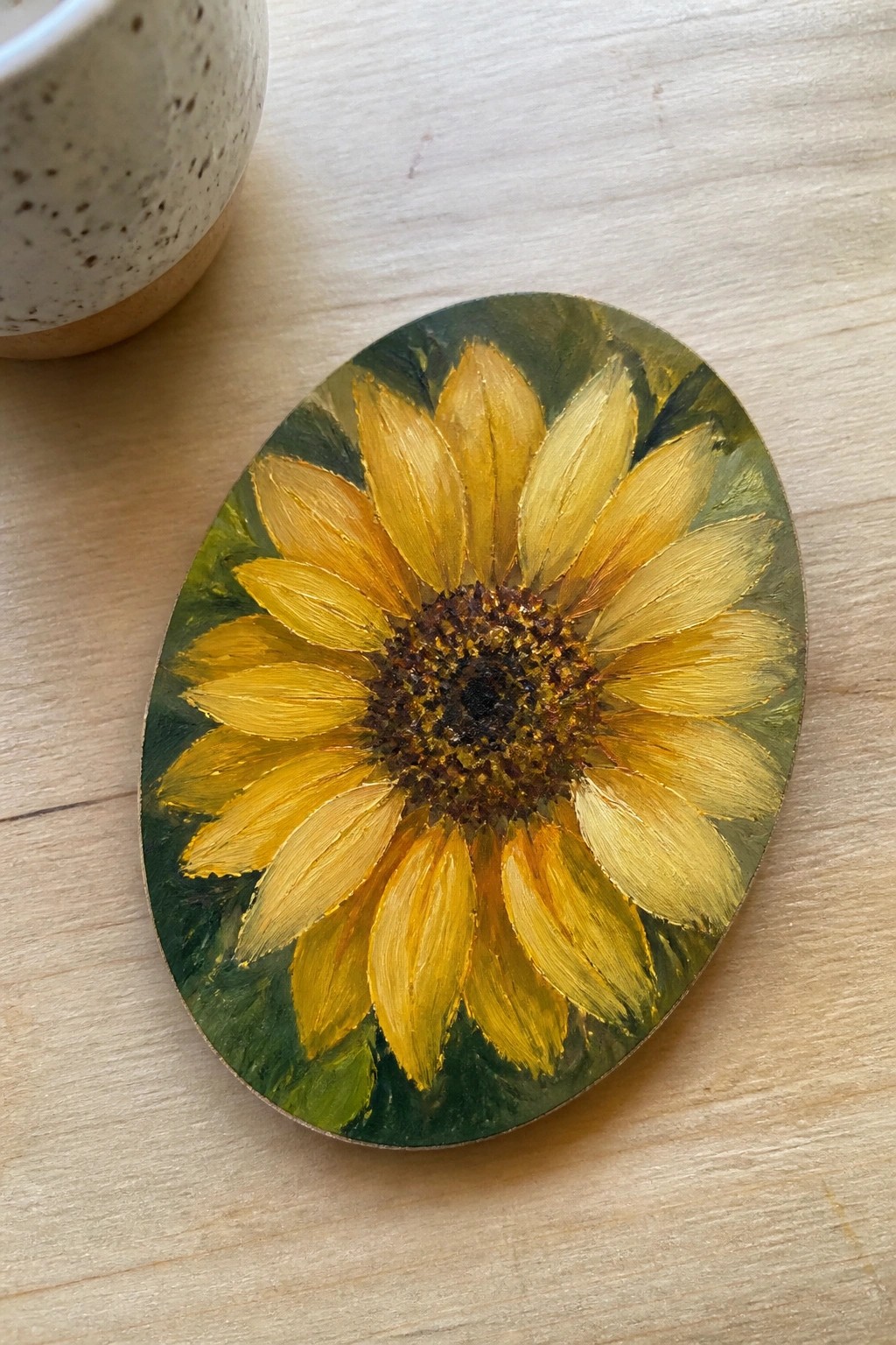Oval yellow sunflower painting on wooden table beside speckled ceramic mug