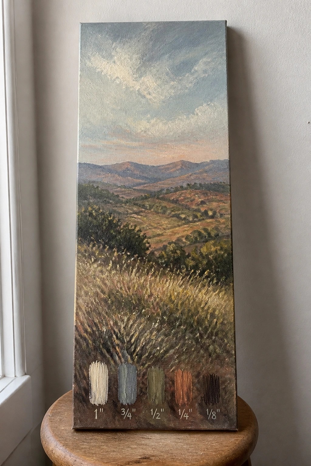 Tall vertical landscape painting of hills with labeled paint swatches on canvas stool.