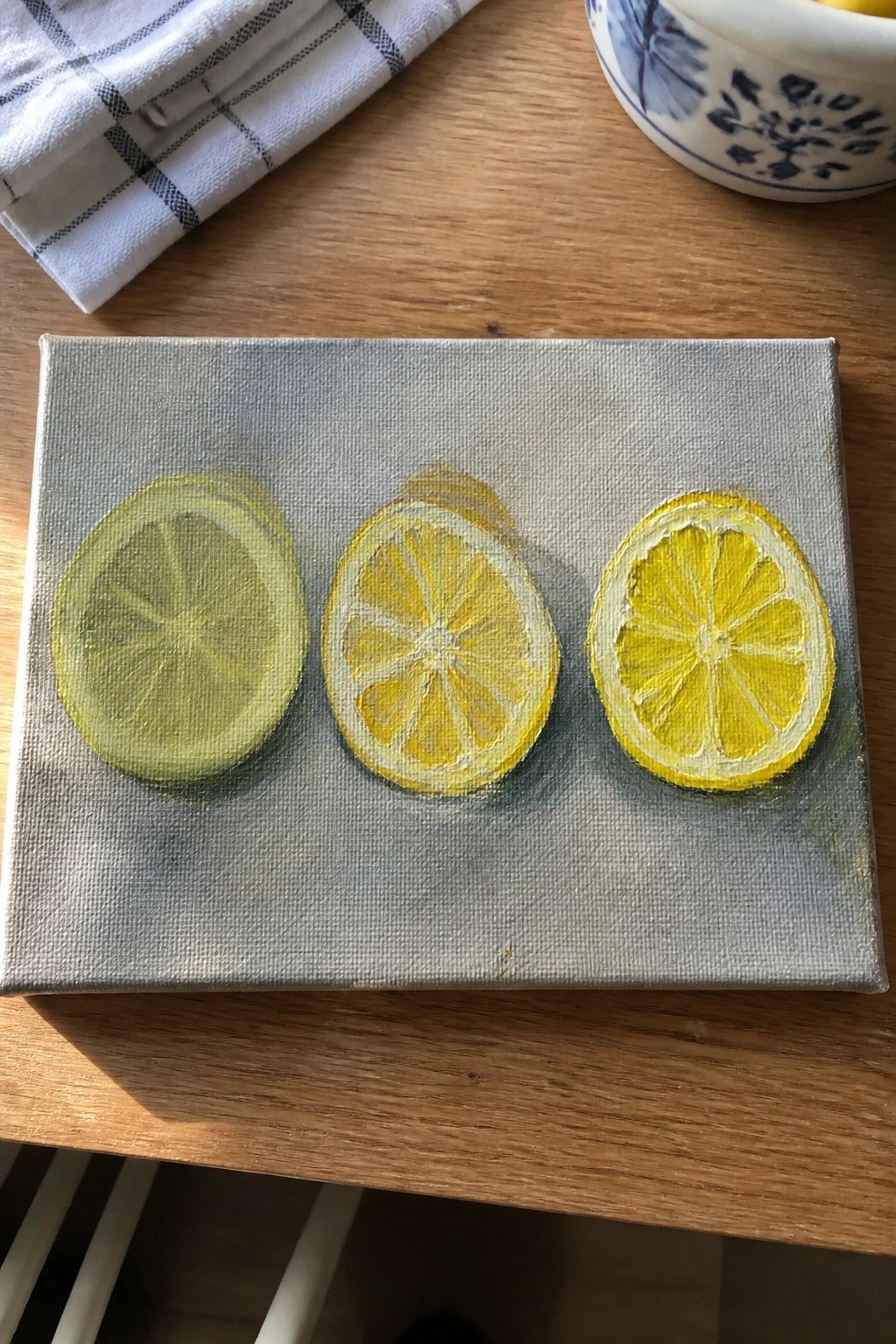 Three lemon slices painted yellow-green on small gray canvas atop wooden table