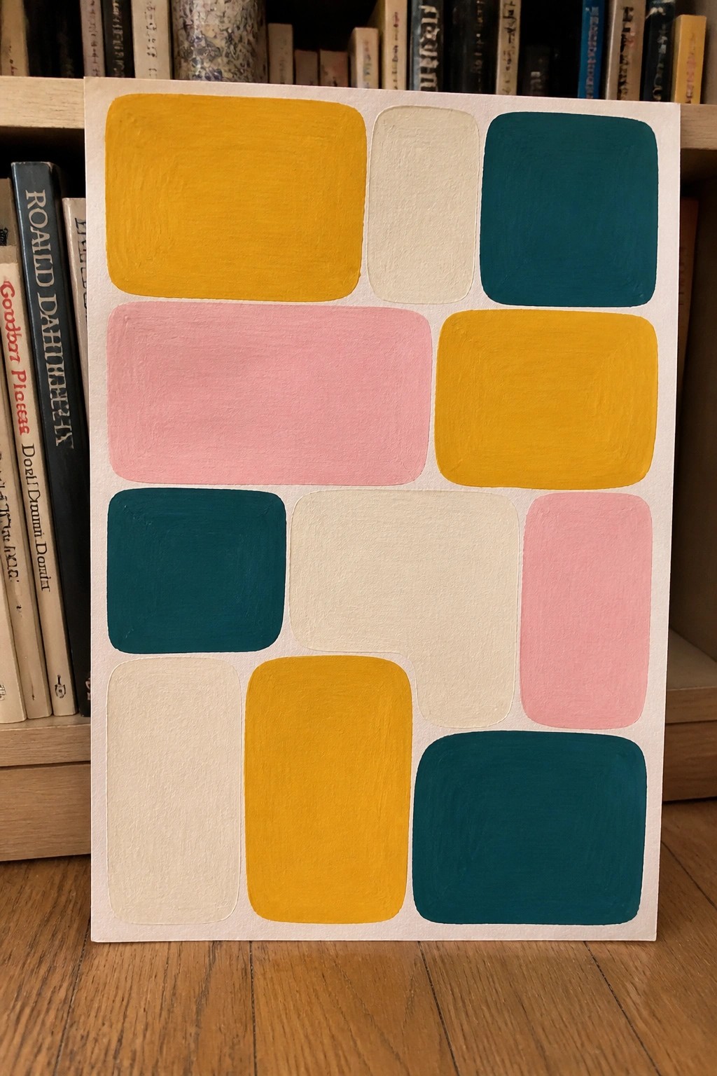 Abstract geometric painting with rounded shapes in yellow, pink, teal, and beige against books