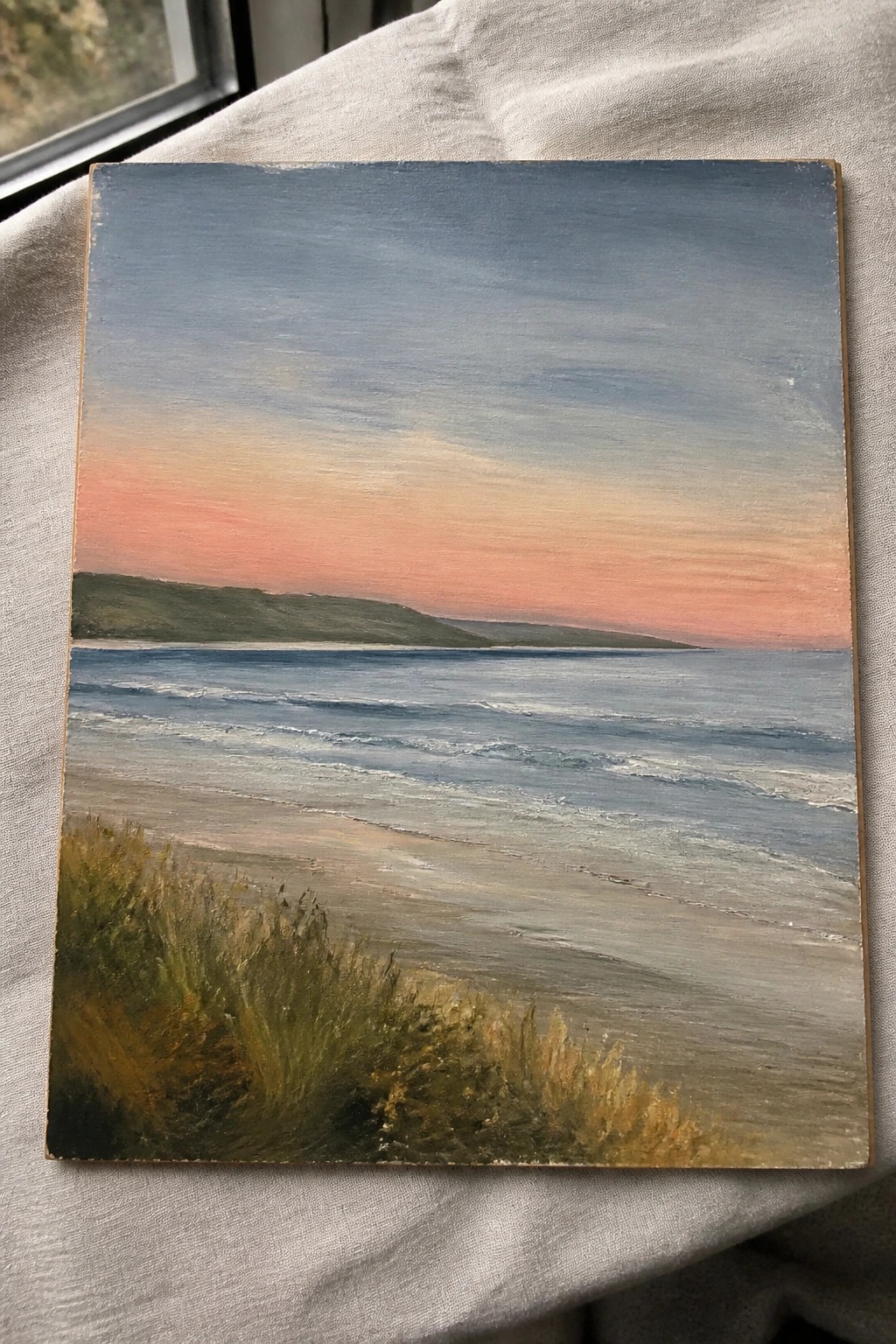 An oil painting of a beach at sunset with a pink and blue sky, waves on the water, a distant green hill, and grass in the foreground.