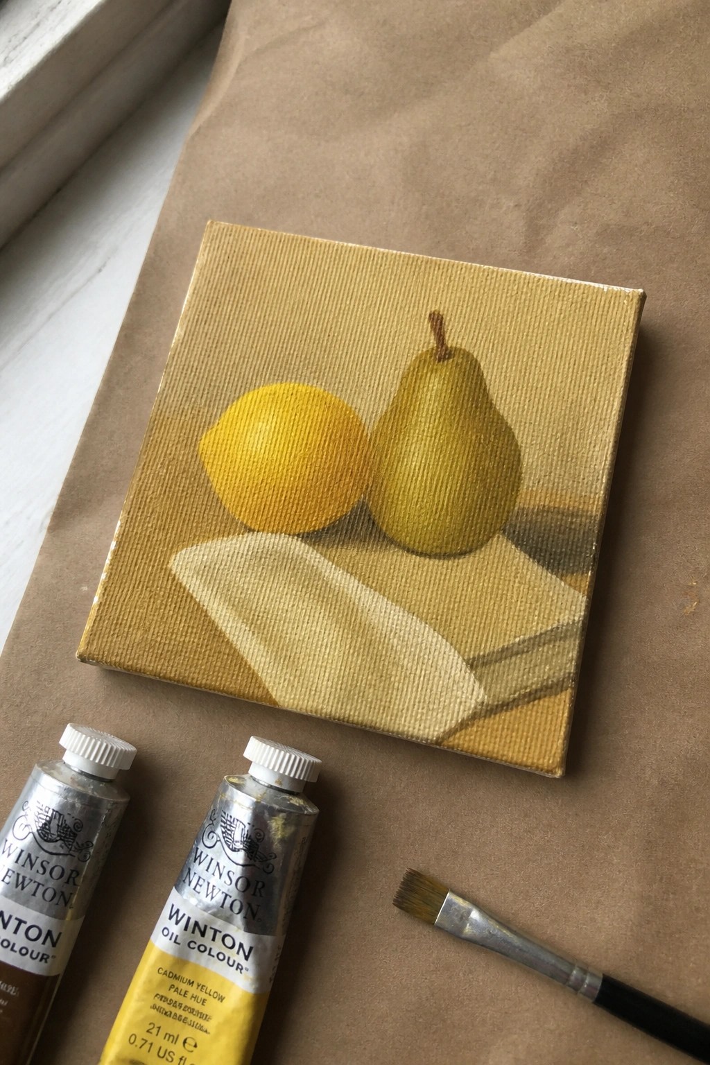 Oil painting of yellow lemon and pear beside paint tubes and brush on paper.