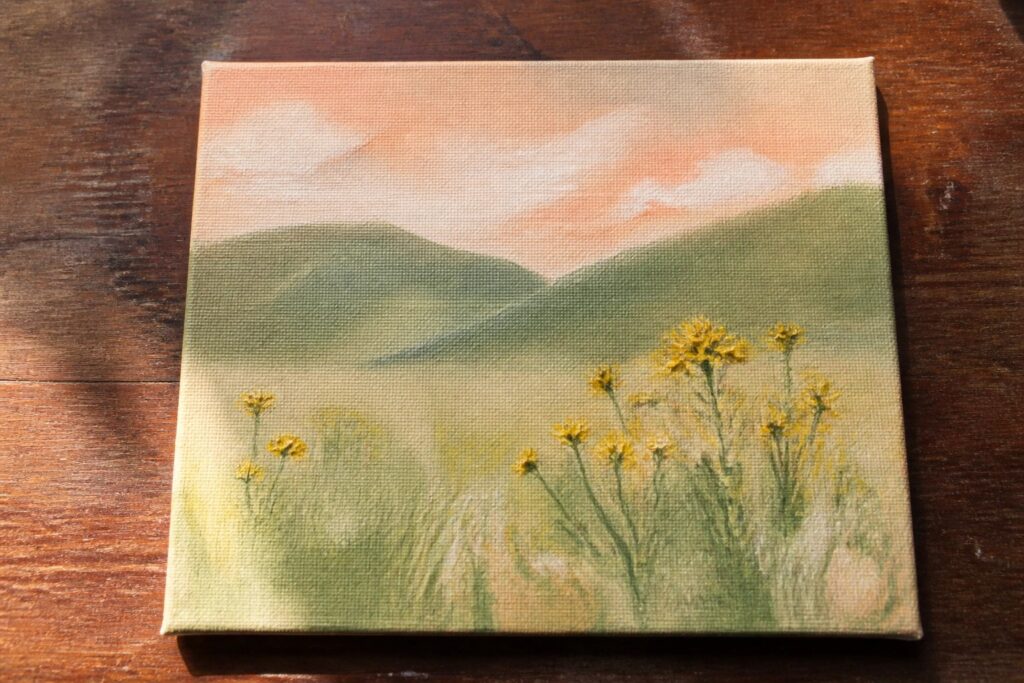 An oil painting of green rolling hills with yellow wildflowers in the foreground under a soft orange and cream sky on a canvas resting on wood.