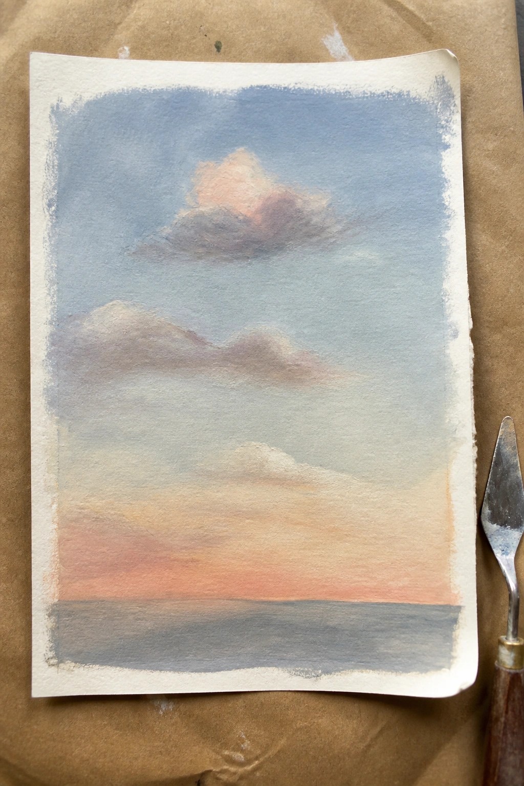 Watercolor sky study with blue-pink clouds over orange horizon, palette knife beside it