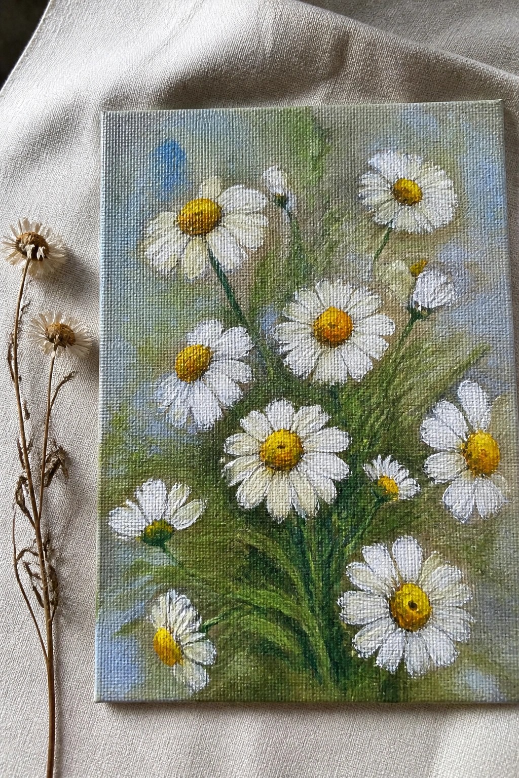Canvas painting of white daisies with yellow centers on white fabric beside dried flowers.