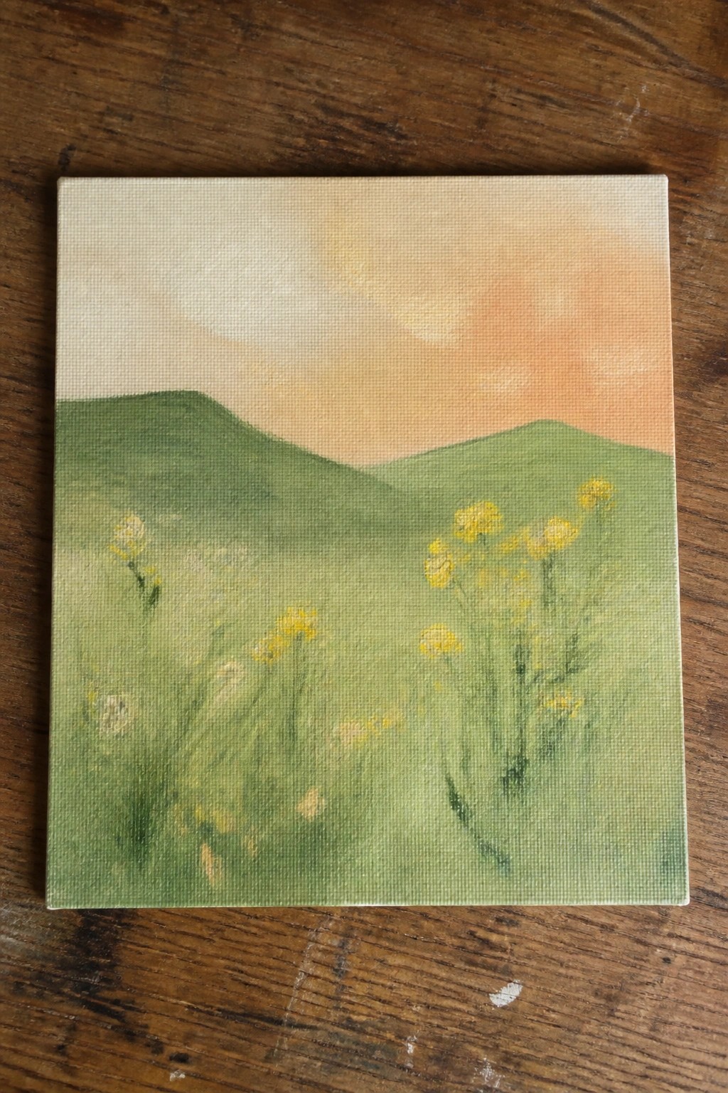 An oil painting of green rolling hills with yellow wildflowers in the foreground under a soft orange and cream sky on a canvas resting on wood.