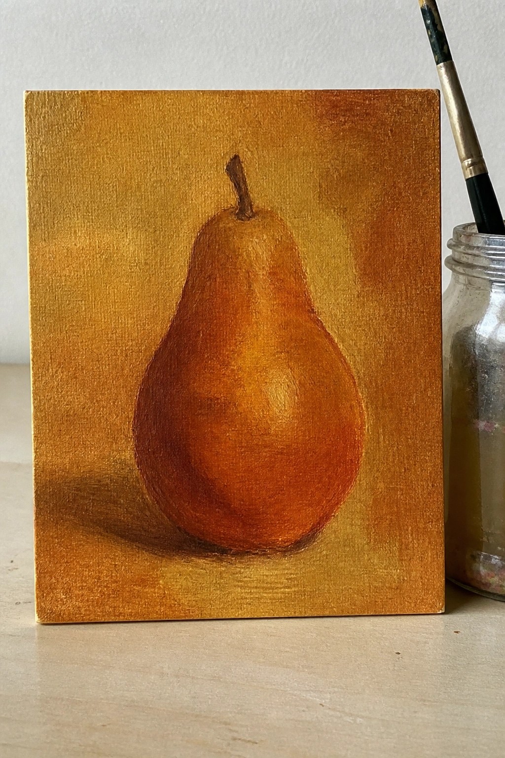 Oil painting of brown pear on golden canvas beside paintbrush in jar