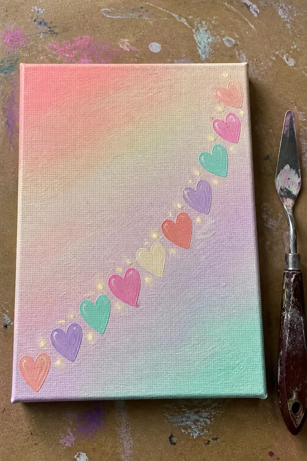 Canvas with rainbow gradient and curved colorful hearts beside palette knife on paint table