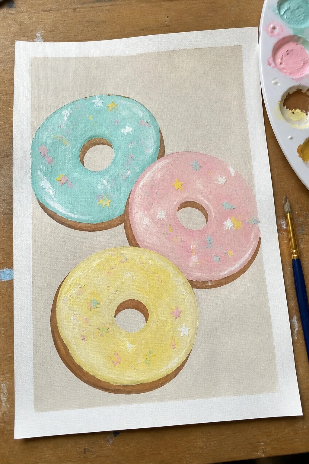 Three pastel-frosted donuts with star sprinkles painted on canvas beside palette and brush