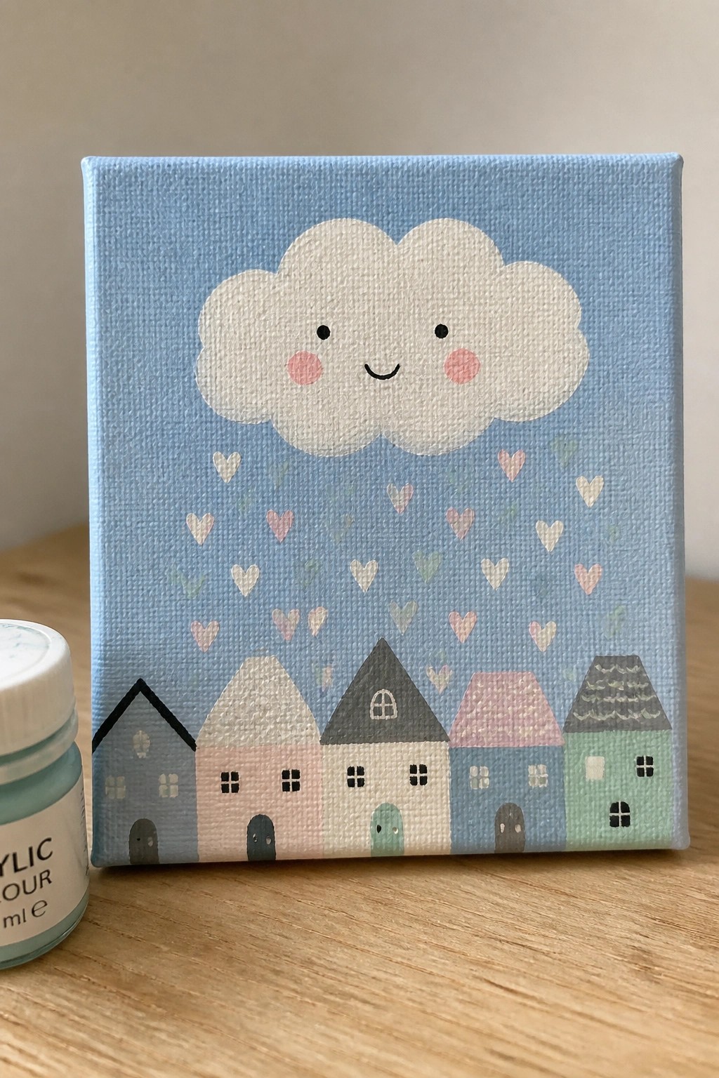 Cute canvas painting of smiling cloud raining hearts above row of colorful houses, paint jar nearby.