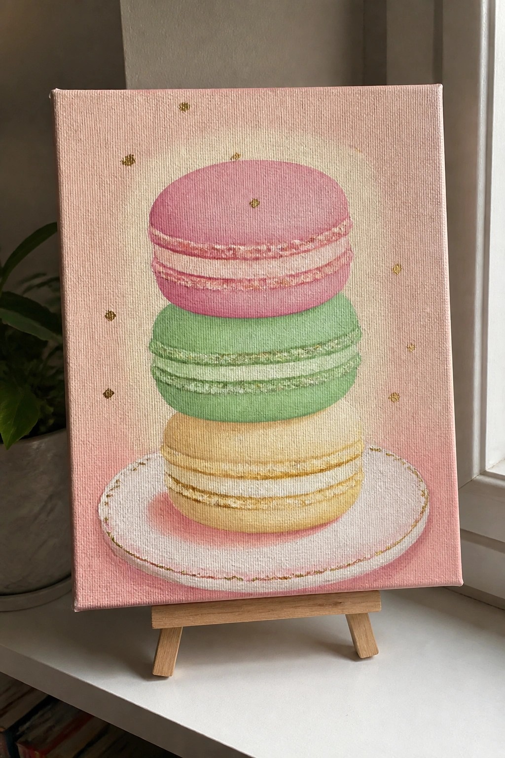 Stacked pink, green, and yellow macarons on gold-rimmed plate, pink canvas with gold dots on easel