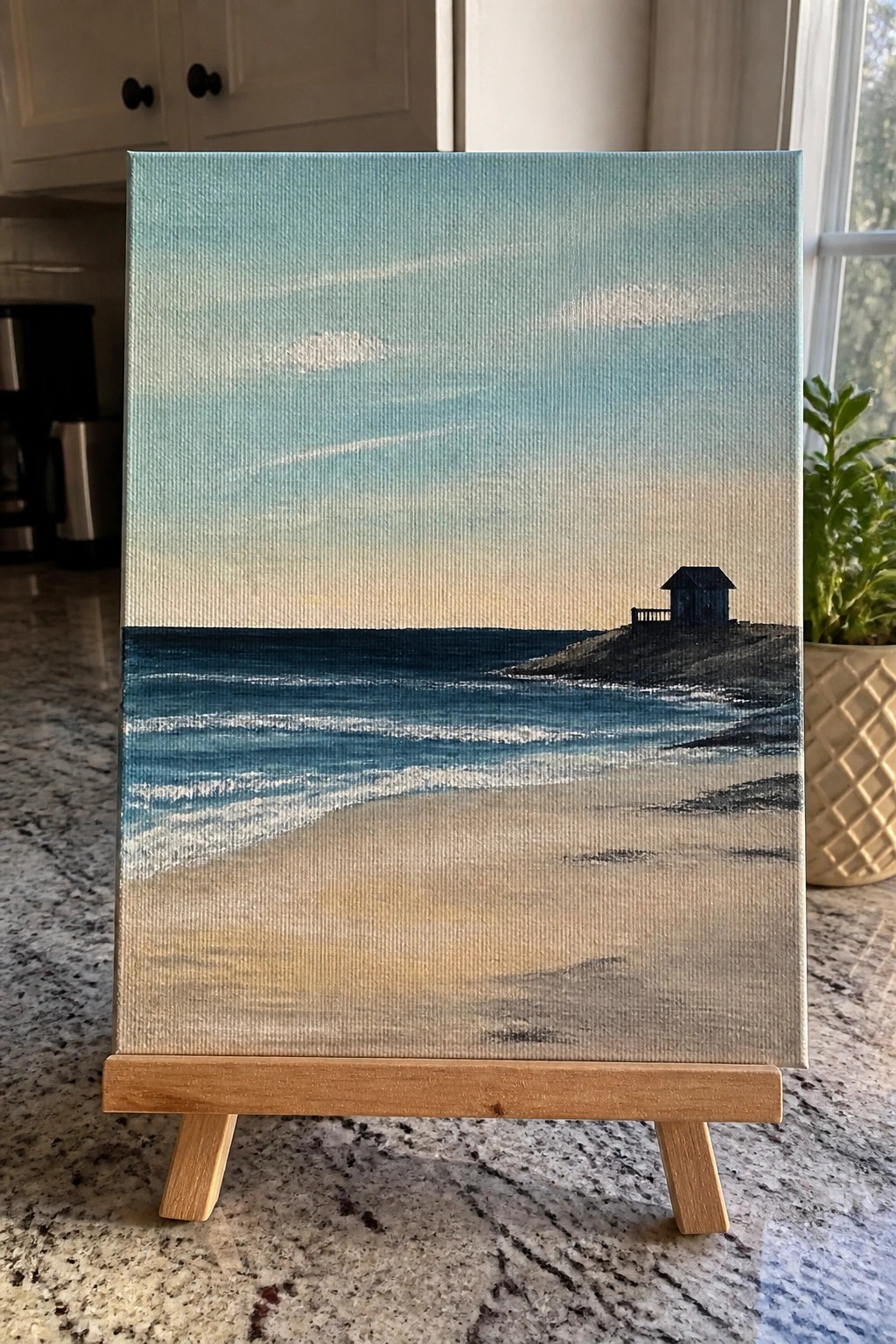 Seascape painting on wooden easel showing beach, waves, and small dark house under blue sky.