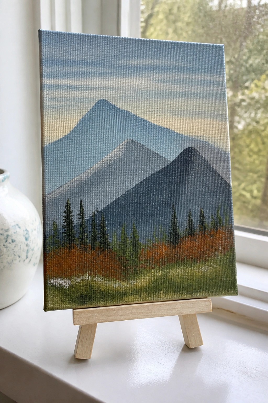 Small canvas painting of blue mountains and pine trees on wooden easel by window