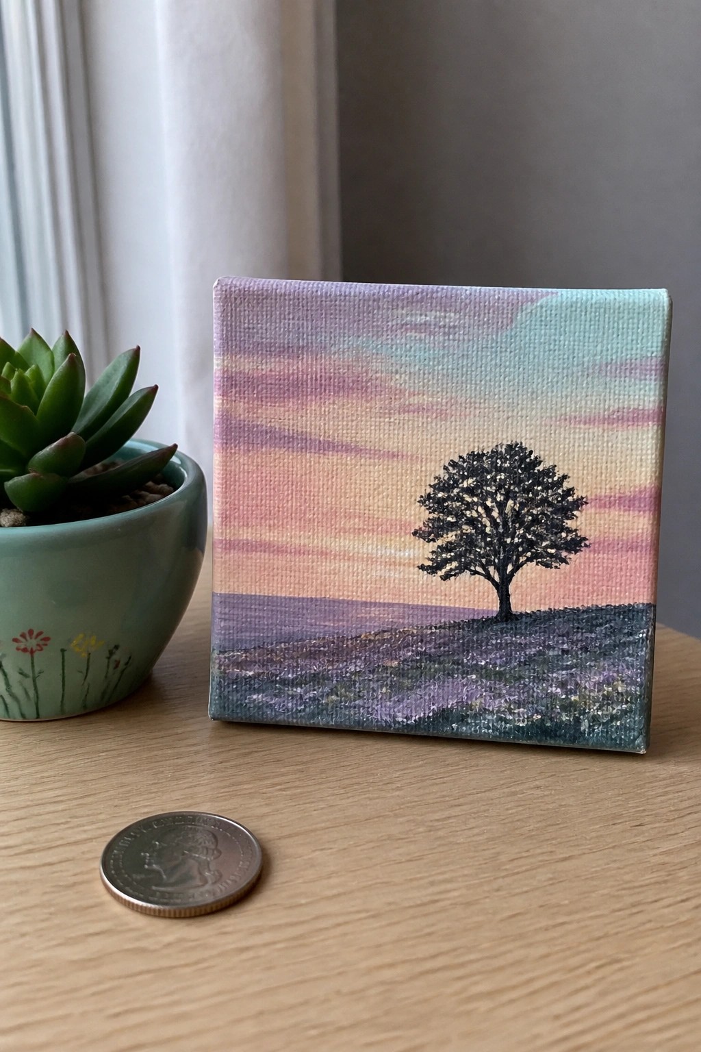 Small painting of lone tree at colorful sunset beside potted succulent and coin on table