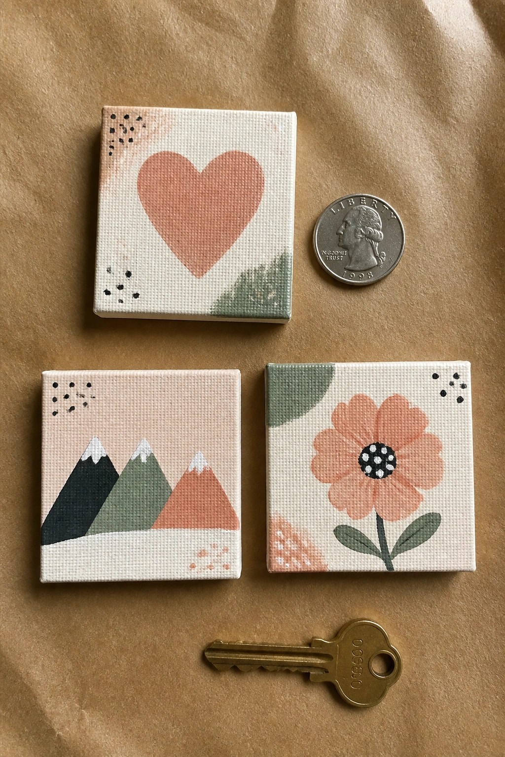 Three miniature canvases with heart, mountains, and flower beside quarter and key.