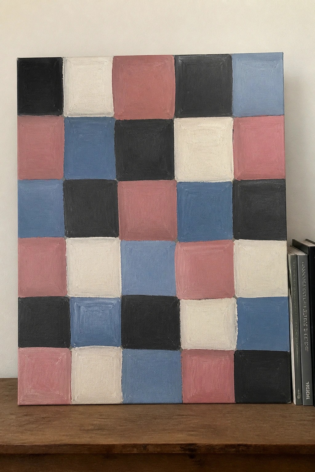 A square oil painting on canvas featuring a grid of colored squares in black, white, pink, and blue tones resting on a wooden surface.