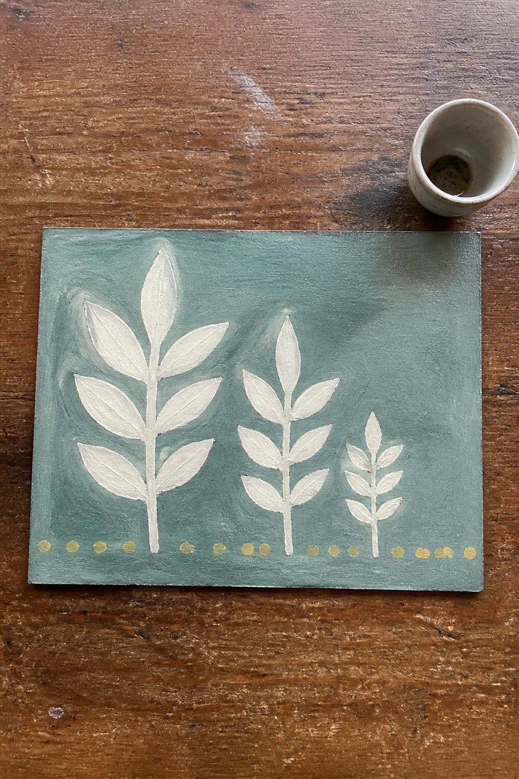 Teal canvas with three white leaf plants, gold dots, beside cup on wood table