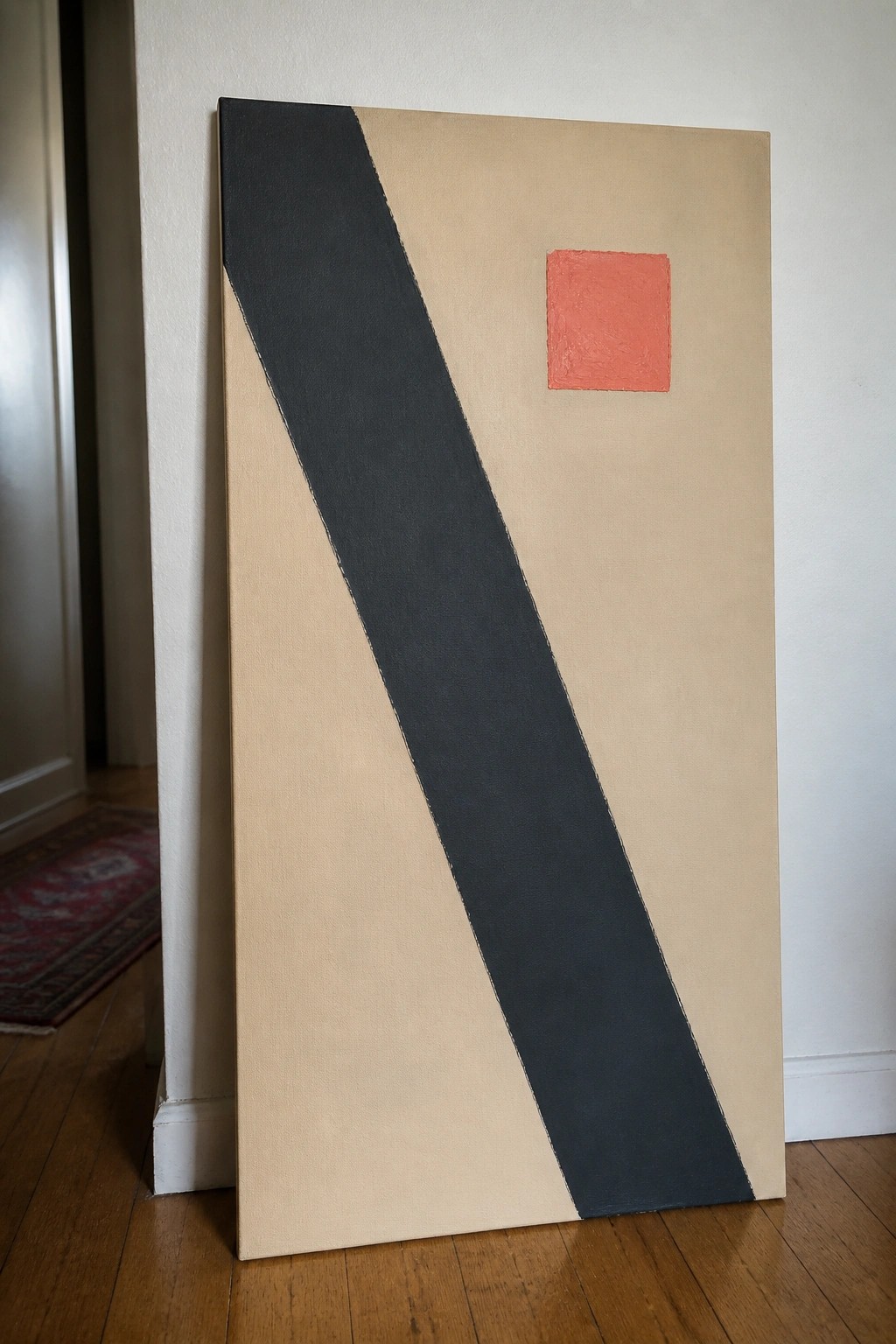 Abstract painting with beige background, diagonal black stripe, and red square leaning on wall