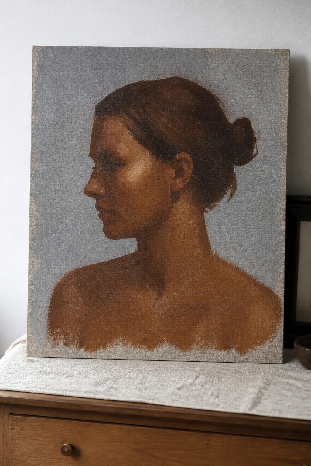 Profile portrait painting of bare-shouldered woman with hair in bun on wooden surface.