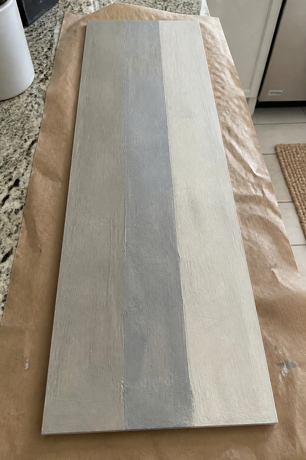 Wooden board with three vertical stripes of beige, gray, and white paint on brown paper.
