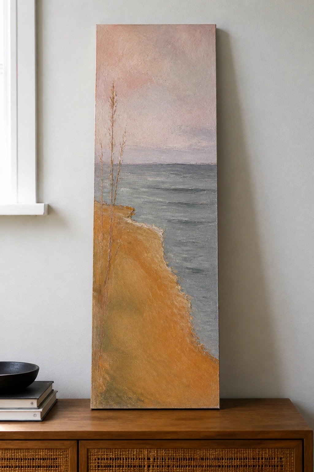 Tall landscape painting of orange cliff and gray sea on wooden cabinet with books and bowl