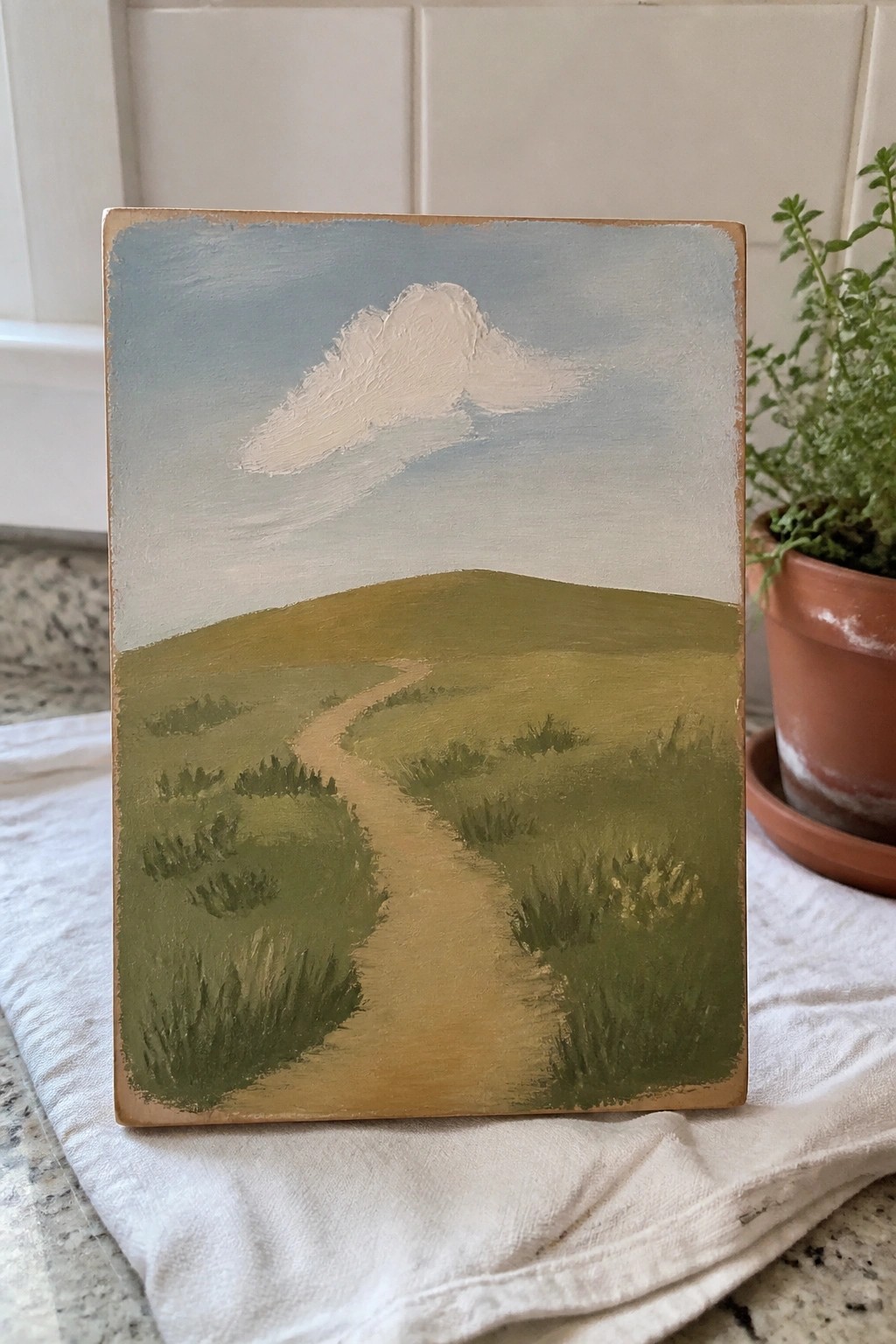 An oil painting of a winding path through green hills with a white cloud in a blue sky, displayed on a cloth next to a potted plant.
