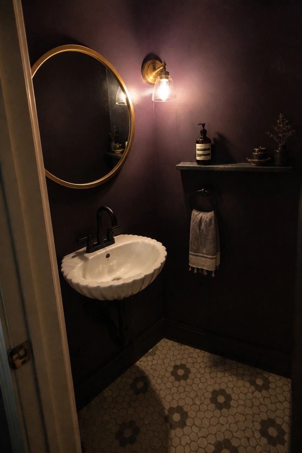 A small bathroom with deep purple walls, a white scalloped sink, and warm brass lighting.