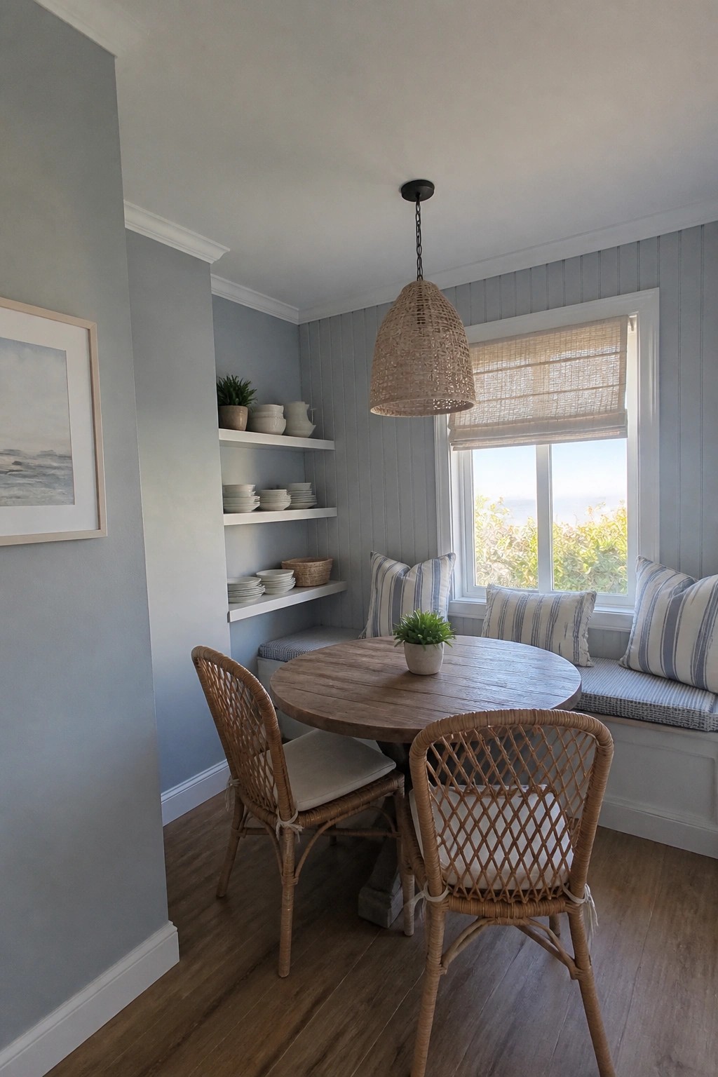 Coastal dining nook with round wooden table, rattan chairs, striped bench, and ocean view.