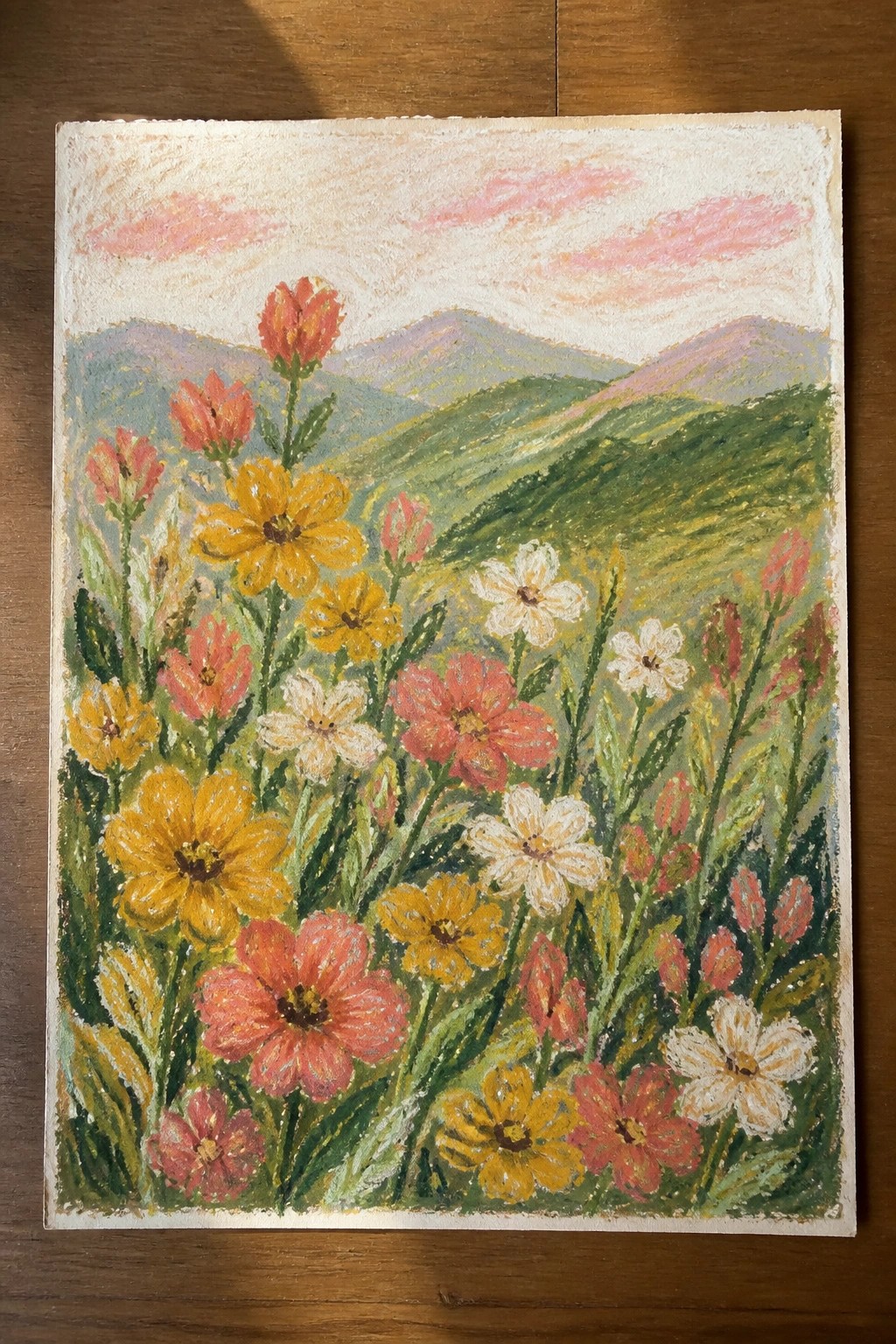 Vibrant wildflowers in yellow, pink, and white bloom before layered green mountains under a pink sky.