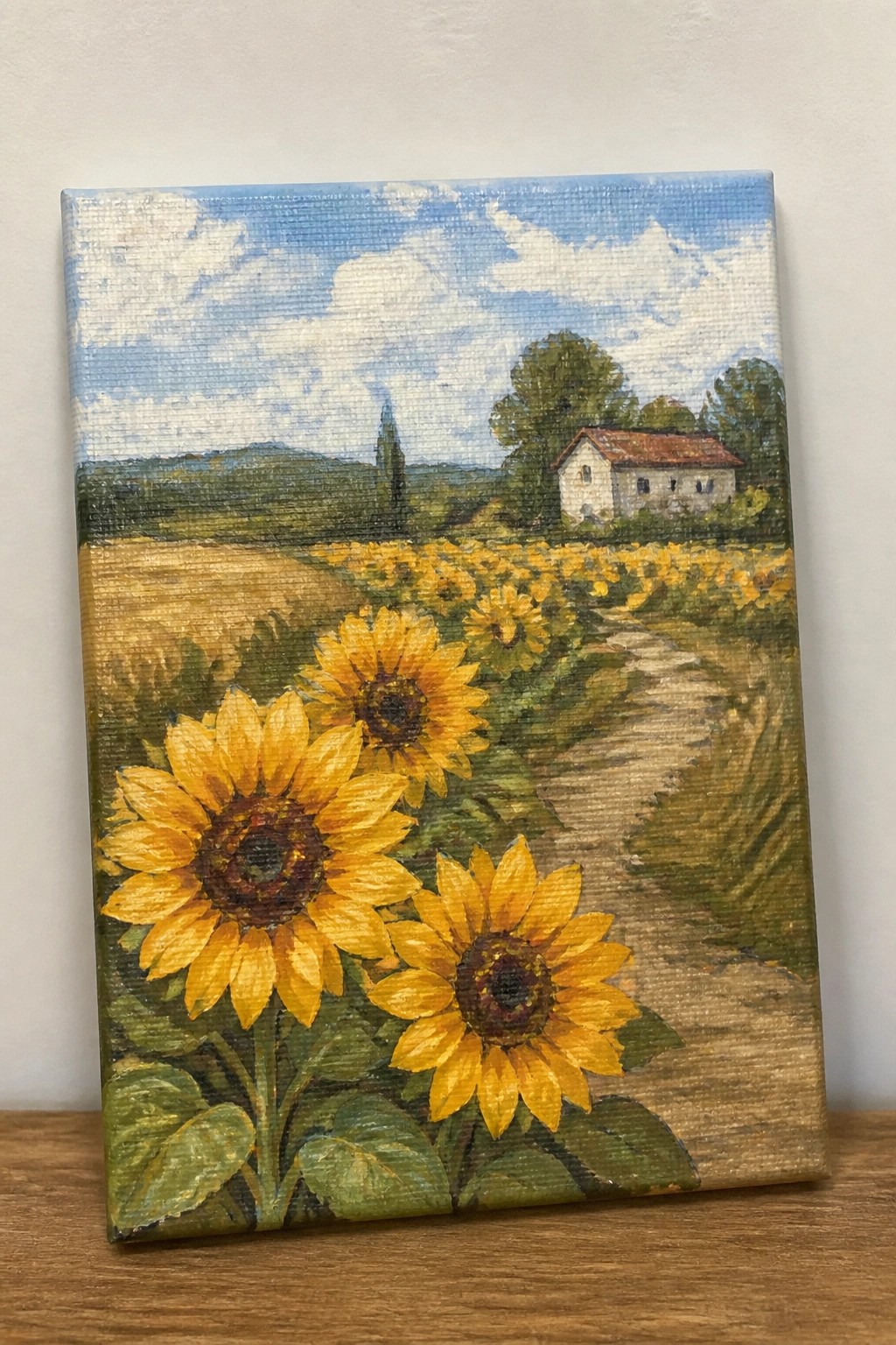 Oil painting of sunflowers by a winding path to a white house under blue skies.